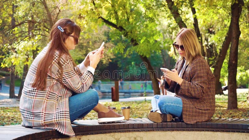 Chicas elegantes sentadas en el parque y usando teléfonos inteligentes