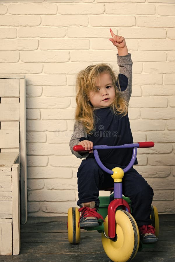 Child Childhood Children Happiness Concept. Happy childhood concept. Baby cyclist with long blond hair on white brick wall. Boy riding bicycle in room. Playing and having fun. Child rising index finger on tricycle.