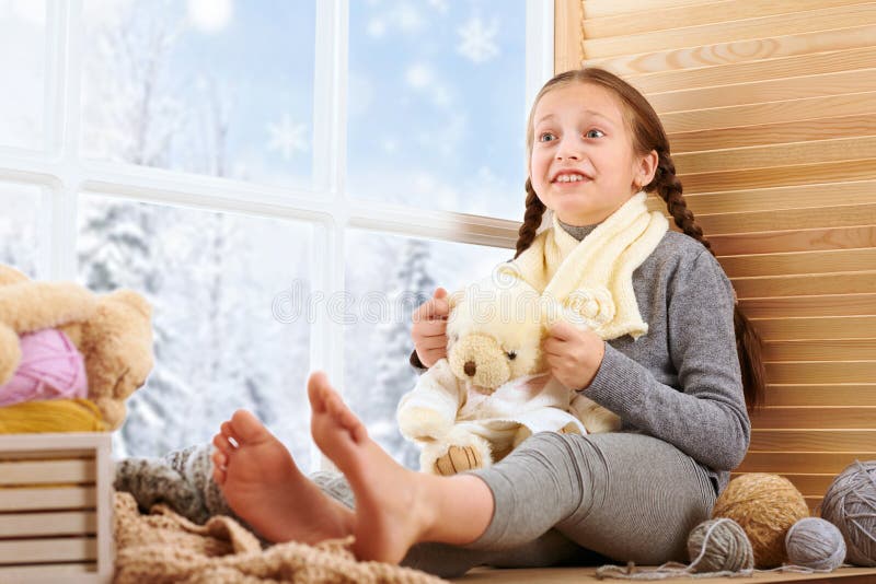 Child girl is sitting on a window sill and playing with bear toy. Beautiful view outside the window - sunny day in winter and snow
