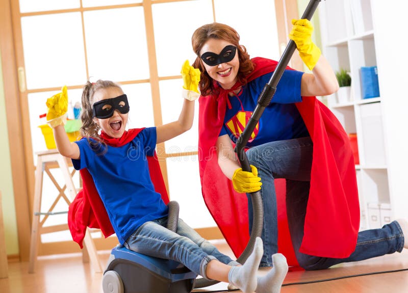 Child and mother dressed as superheroes using vacuum cleaner in room. Family middle-aged woman and daughter have a fun