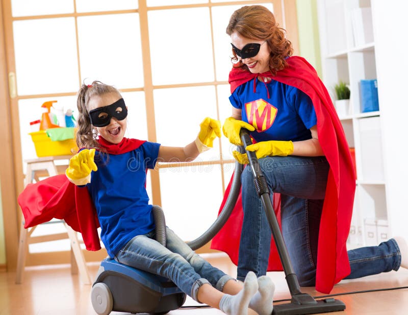 Child and mother dressed as superheroes using vacuum cleaner in room. Family middle-aged woman and daughter have a fun