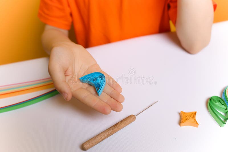 Child in an orange T-shirt shows twisted paper strips in the quilling technique. the boy prepared the blanks for gluing them to stock photos