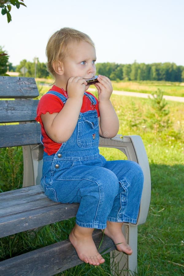 A toddler girl dressed in a red shirt and denim overalls and with bare feet sitting on a bench playing the harmonica. A toddler girl dressed in a red shirt and denim overalls and with bare feet sitting on a bench playing the harmonica.