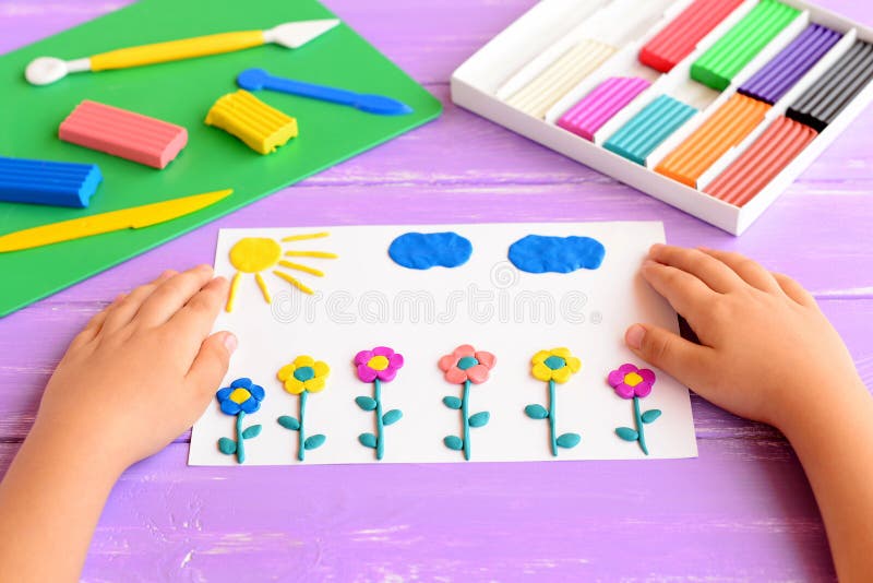 Child shows a card with plasticine flowers, sun and clouds. Supplies for children art crafts on wooden table. Modeling clay craft