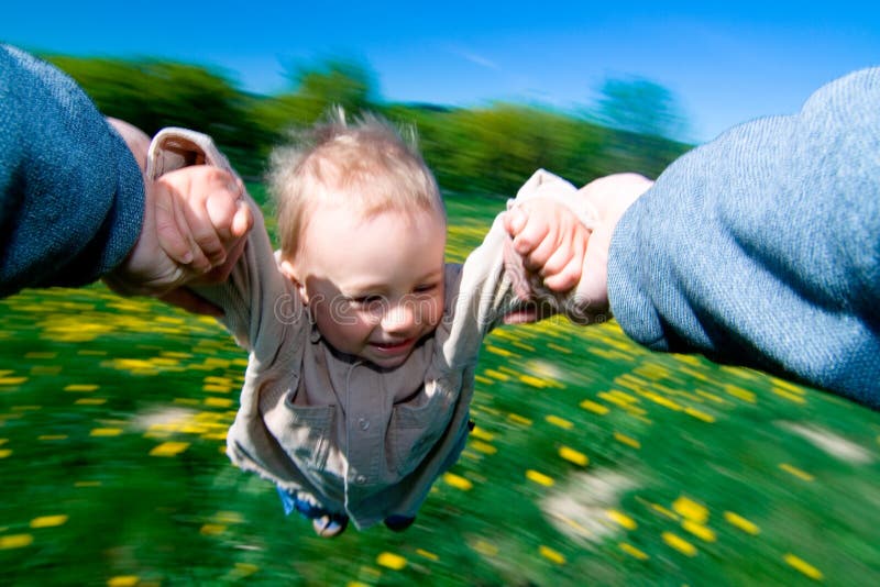 Bambina o bambino, di essere oscillato intorno in cerchi da ogni braccio da un adulto che indossa maglione blu, verde erba in background, movimento offuscata.