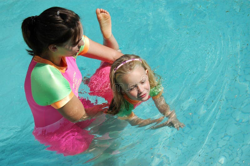 Una bella bionda caucasica bianca piccola ragazza, con felice espressione emozionante nel suo bel volto che indossa un costume coloratissimo avere un grande divertimento con l'apprendimento di nuotare nell'acqua di una piscina di essere aiutato dal suo femmina insegnante di sport all'aria aperta, nel sole.