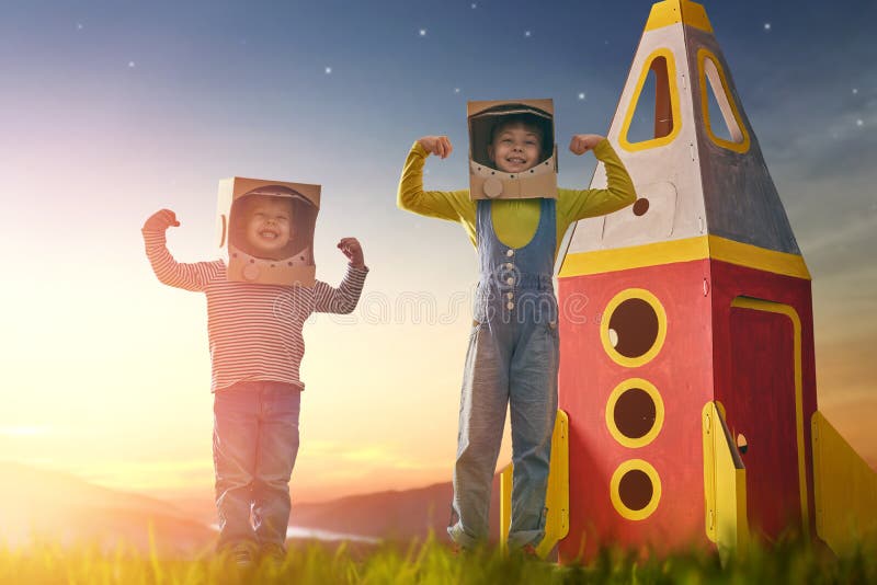 Children in astronauts costumes with toy rocket playing and dreaming of becoming a spacemen. Portrait of funny kids on a background of sunset star sky on nature. Family friends games outdoors.