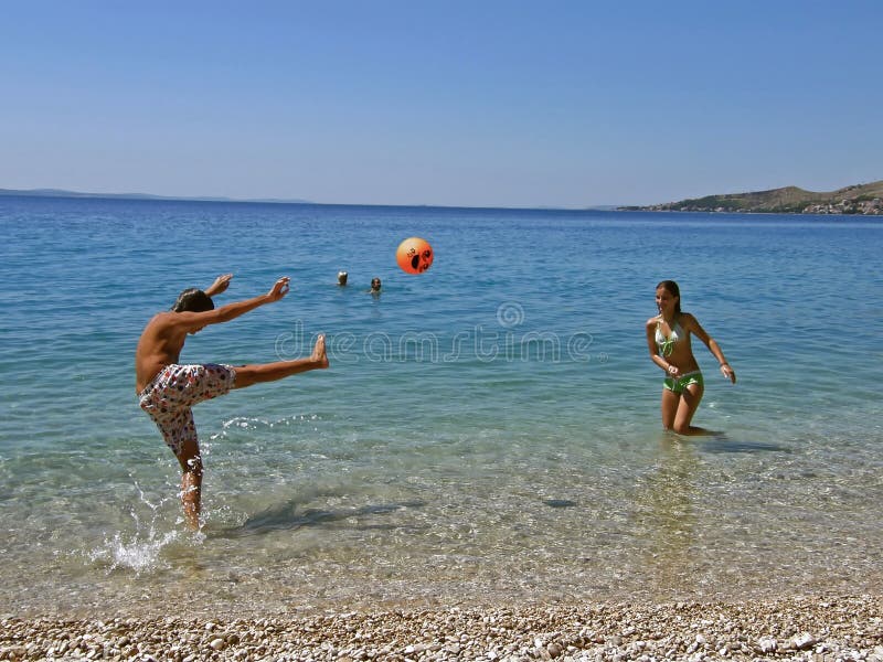 Boy and girl (best friends) play with a ball - gave a fun - games in (boy kicking a orange ball) shallow at crystal clear Adriatic sea - Croatia - Dalmatia at sunny day. Horizontal color photo. Boy and girl (best friends) play with a ball - gave a fun - games in (boy kicking a orange ball) shallow at crystal clear Adriatic sea - Croatia - Dalmatia at sunny day. Horizontal color photo.