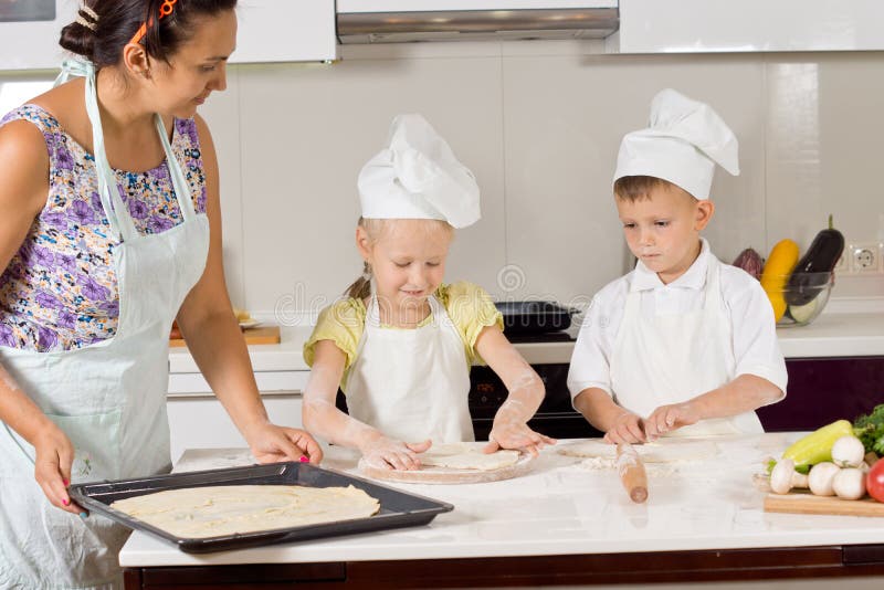 Two cute children wearing chef uniform while helping their mother to prepare the dough for baking a homemade pie, in the kitchen. Two cute children wearing chef uniform while helping their mother to prepare the dough for baking a homemade pie, in the kitchen