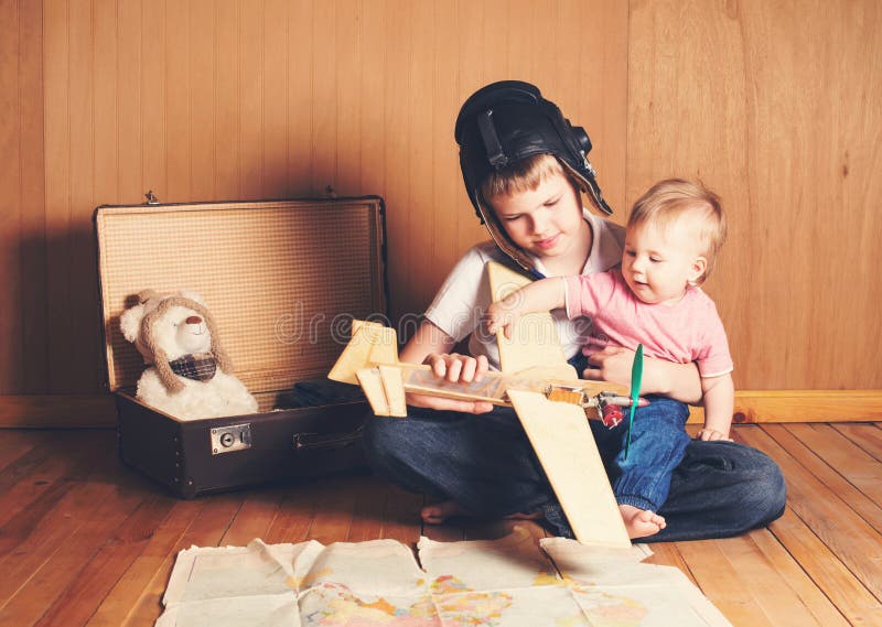 Children playing. Boy in pilot helmet with plane model and suitcase, dreaming of adventure. Journey. Brother holding his little b stock photo Children playing. Boy in pilot helmet with plane model and suitcase, dreaming of adventure. Journey. Brother holding his little b stock photo