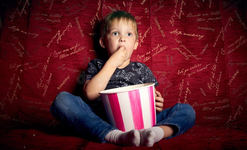 Children`s cinema: A little boy, a preschooler, watch a movie at home on a big red sofa and eat popcorn from a big
