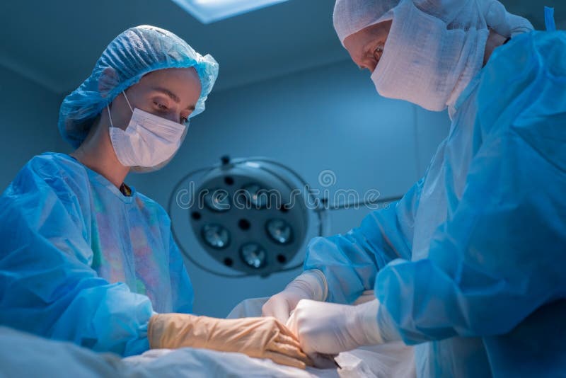 Children`s surgeons perform urological operations. A men and a women in a mask and a blue sterile coat in the operating room. A team of surgeons is conducting an operation. Treatment of inguinal hernia, testicular edema, testicular disease, varicocele, cryptorchidism. Focus on the warm light of the lamp, the background is illuminated by cold blue light. bottom view. Children`s surgeons perform urological operations. A men and a women in a mask and a blue sterile coat in the operating room. A team of surgeons is conducting an operation. Treatment of inguinal hernia, testicular edema, testicular disease, varicocele, cryptorchidism. Focus on the warm light of the lamp, the background is illuminated by cold blue light. bottom view