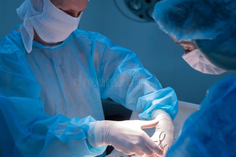 Children`s surgeons perform urological operations. A men and a women in a mask and a blue sterile coat in the operating room. A team of surgeons is conducting an operation. Treatment of inguinal hernia, testicular edema, testicular disease, varicocele, cryptorchidism. Focus on the warm light of the lamp, the background is illuminated by cold blue light. Children`s surgeons perform urological operations. A men and a women in a mask and a blue sterile coat in the operating room. A team of surgeons is conducting an operation. Treatment of inguinal hernia, testicular edema, testicular disease, varicocele, cryptorchidism. Focus on the warm light of the lamp, the background is illuminated by cold blue light