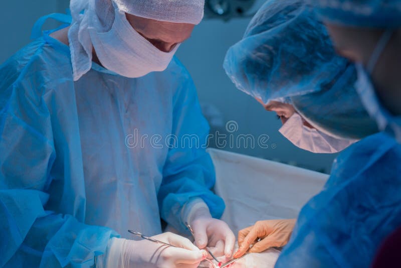 Children`s surgeons perform urological operations. A men and a women in a mask and a blue sterile coat in the operating room. A team of surgeons is conducting an operation. Treatment of inguinal hernia, testicular edema, testicular disease, varicocele, cryptorchidism. Focus on the warm light of the lamp, the background is illuminated by cold blue light. Children`s surgeons perform urological operations. A men and a women in a mask and a blue sterile coat in the operating room. A team of surgeons is conducting an operation. Treatment of inguinal hernia, testicular edema, testicular disease, varicocele, cryptorchidism. Focus on the warm light of the lamp, the background is illuminated by cold blue light
