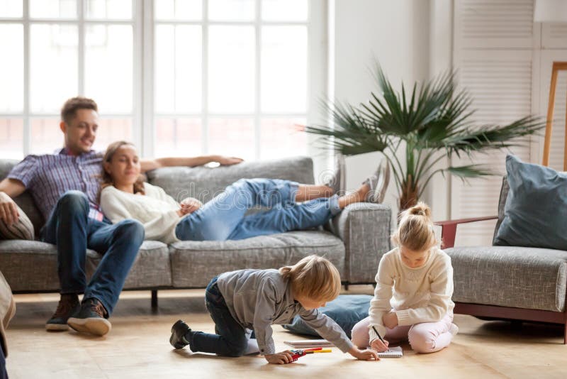 Children sister and brother playing drawing together on floor while young parents relaxing at home on sofa, little boy girl having fun, friendship between siblings, family leisure time in living room. Children sister and brother playing drawing together on floor while young parents relaxing at home on sofa, little boy girl having fun, friendship between siblings, family leisure time in living room