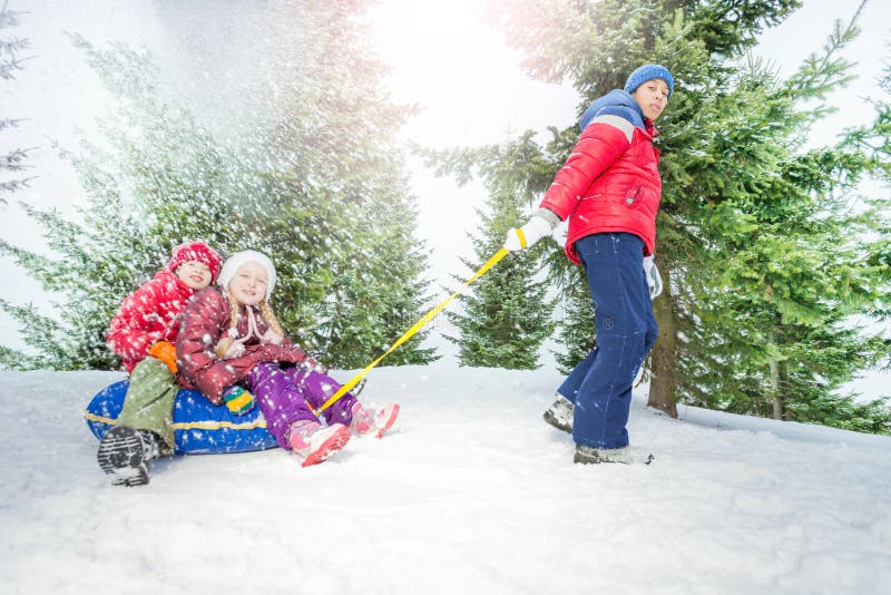 Children sitting on snow tube and other boy pulling them in winter during day in the fir tree forest. Children sitting on snow tube and other boy pulling them in winter during day in the fir tree forest