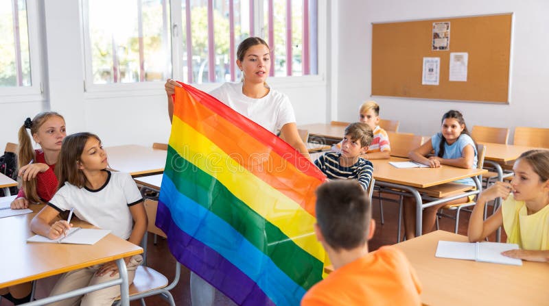Children sitting in classroom and listening to female teacher. She holding rainbow flag in hands and talking about