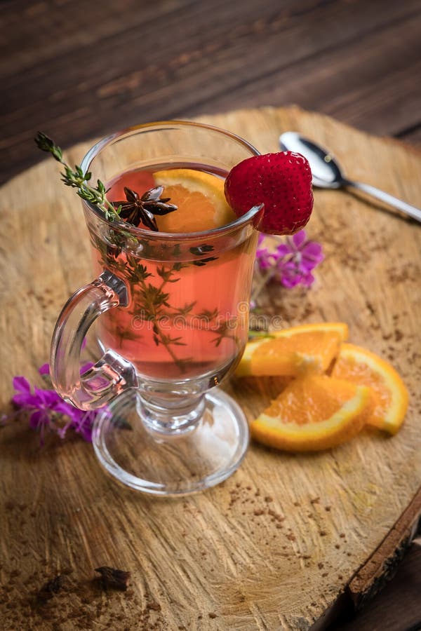 Chili cinnamon bourbon hot toddy in transparent glass on wooden cross section of trunk tree decorated with mint, strawberry and orange. Chili cinnamon bourbon hot toddy in transparent glass on wooden cross section of trunk tree decorated with mint, strawberry and orange