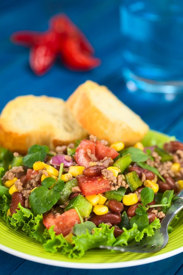 Chili con carne salad made of mincemeat, kidney beans, watercress, green bell pepper, tomato, sweet corn and red onions served on lettuce on a plate with fork and baguette slices (Selective Focus, Focus in the middle of the salad). Chili con carne salad made of mincemeat, kidney beans, watercress, green bell pepper, tomato, sweet corn and red onions served on lettuce on a plate with fork and baguette slices (Selective Focus, Focus in the middle of the salad)