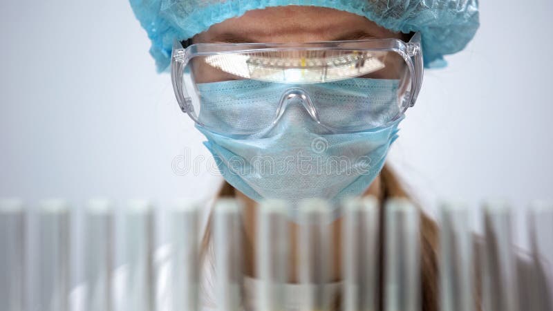 Chimico femminile nella maschera protettiva che esamina le provette, ricerca biologica fotografia stock Chimico femminile nella maschera protettiva che esamina le provette, ricerca biologica fotografia stock