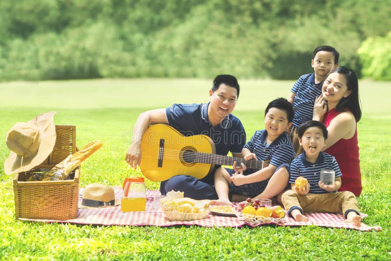 Chinese family smiling at the camera while playing a guitar and singing together in the park. Chinese family smiling at the camera while playing a guitar and singing together in the park