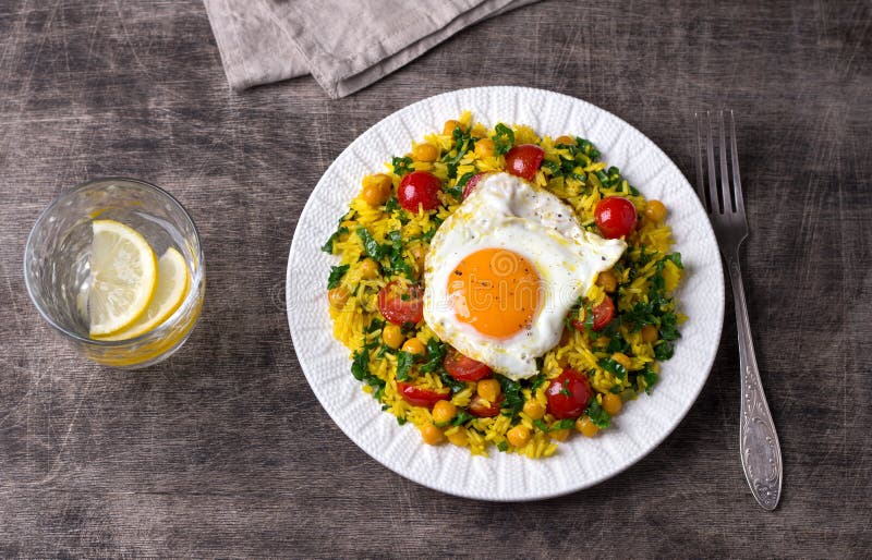 Chinese fried rice with turmeric, chickpeas, tomatoes, kale and with fried eggs on white ceramic plate on dark wooden background