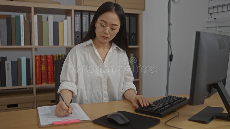Chinese woman taking notes at her workplace in an office environment, surrounded by bookshelves and working with documents on her royalty free stock photo