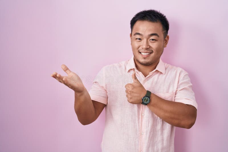 Chinese young man standing over pink background showing palm hand and doing ok gesture with thumbs up, smiling happy and cheerful