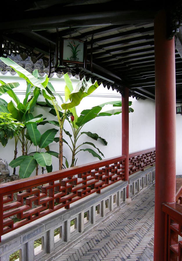 An image of traditional style chinese architecture in an old chinese residence, showing the corridor linking in between buildings. With black wooden beams as support for the roof, red pillars and wood railings carved in oriental patterns. An oriental chinese lantern hangs from the roof of th corridors. The courtyard beside the path is planted with jade green banana trees and landscaped with rocks (for artificial mountains) and grass in a tranquil and meditative horticultural style. Exotic travels photography to asian scenic destinations with art and local culture interest. Asia tourism photo; empty, nobody in picture. An image of traditional style chinese architecture in an old chinese residence, showing the corridor linking in between buildings. With black wooden beams as support for the roof, red pillars and wood railings carved in oriental patterns. An oriental chinese lantern hangs from the roof of th corridors. The courtyard beside the path is planted with jade green banana trees and landscaped with rocks (for artificial mountains) and grass in a tranquil and meditative horticultural style. Exotic travels photography to asian scenic destinations with art and local culture interest. Asia tourism photo; empty, nobody in picture.