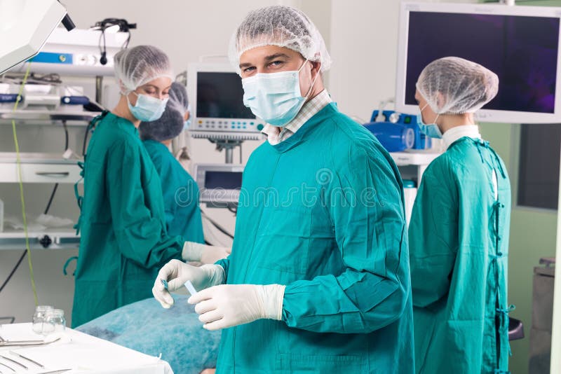 Surgeon with arms crossed looking at camera with colleagues performing in background. Surgeon with arms crossed looking at camera with colleagues performing in background