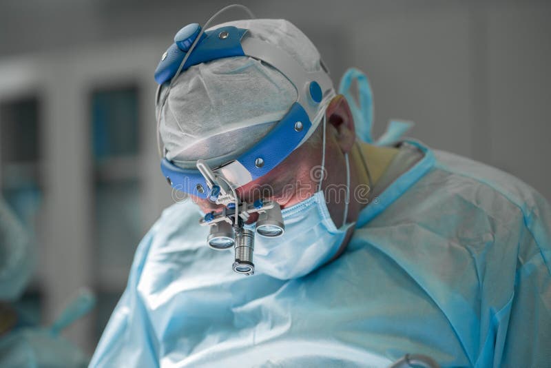 Surgeon performing plastic surgery on breasts in hospital operating room. Surgeon in mask wearing loupes during medical procedure. Breast augmentation, enlargement, enhancement. Surgeon performing plastic surgery on breasts in hospital operating room. Surgeon in mask wearing loupes during medical procedure. Breast augmentation, enlargement, enhancement