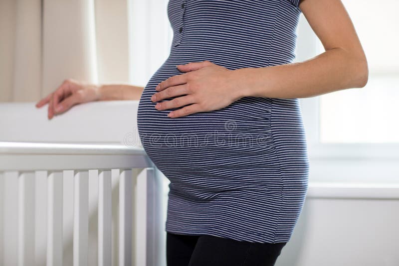 Chiuda su dello stomaco commovente della donna incinta che sta accanto alla culla