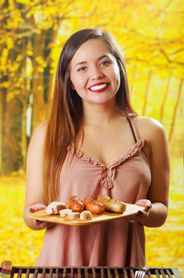 Chiuda su di bella tenuta sorridente della giovane donna in sue mani le salsiccie arrostite sul tagliere di legno, BBQ in immagini stock Chiuda su di bella tenuta sorridente della giovane donna in sue mani le salsiccie arrostite sul tagliere di legno, BBQ in immagini stock