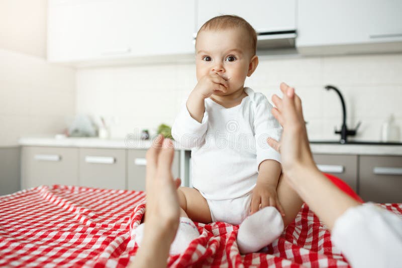 Chiuda su di piccolo neonato allegro con i grandi occhi e mano marroni in bocca che esamina la madre mentre lei che tira le mani fotografia stock libera da diritti Chiuda su di piccolo neonato allegro con i grandi occhi e mano marroni in bocca che esamina la madre mentre lei che tira le mani fotografia stock libera da diritti