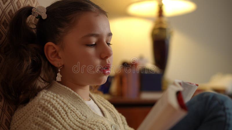 Chiusura di una ragazza seduta su una sedia a dondolo e legge un libro vicino a una lampada da tavolo.