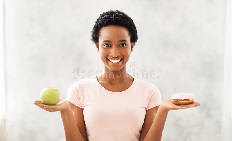 Choice between wholesome and unhealthy food. Happy black woman holding apple and donut, choosing her nutrition