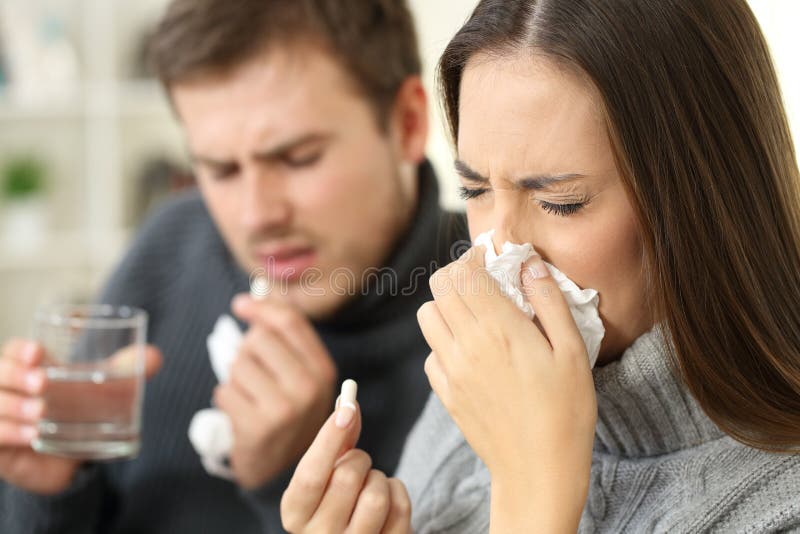 Ill couple wearing jerseys coughing and taking pills sitting on a sofa in the living room in a house interior. Ill couple wearing jerseys coughing and taking pills sitting on a sofa in the living room in a house interior