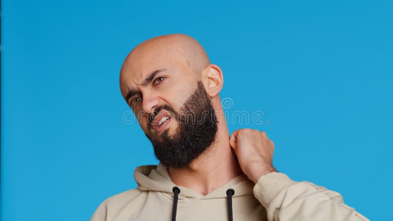 Sick middle eastern guy suffers from neck pain on camera, dealing with sore throat and muscle pressure over blue background. Adult being unwell with spine discomfort in studio. Camera 2. Handheld shot. Sick middle eastern guy suffers from neck pain on camera, dealing with sore throat and muscle pressure over blue background. Adult being unwell with spine discomfort in studio. Camera 2. Handheld shot.