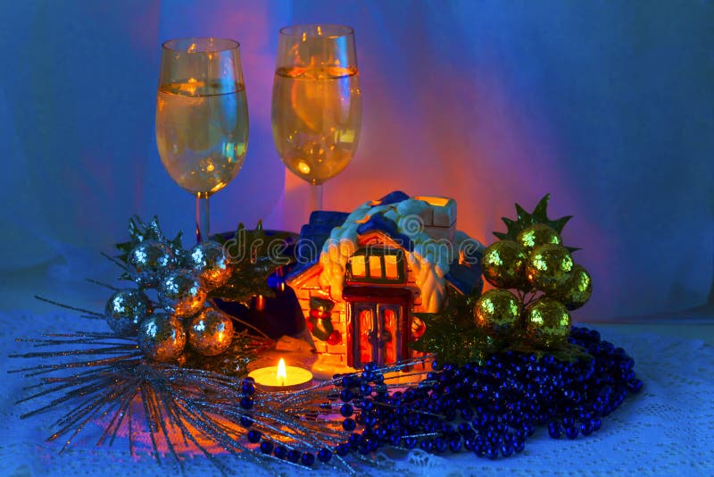 Christmas arrangement with ceramic cabin, candles, wine glasses and Christmas decorations. New Year composition - two glass with sparkling wine glasses, candles, ceramic Christmas house-candlestick, silver decorative branches, blue beads, gold and silver beads, gold and silver in the background, blue and orange tone of the image. Fabulous Christmas picture.