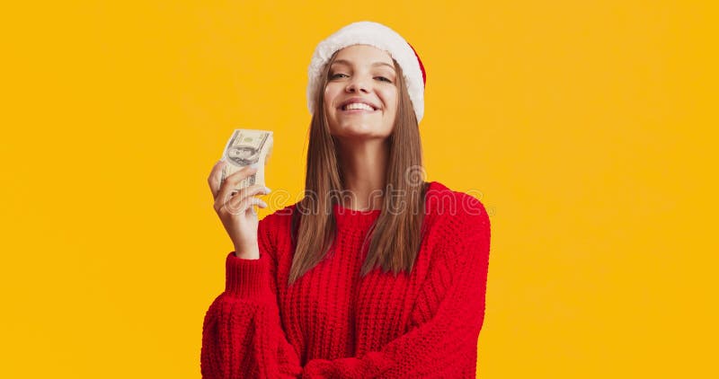 Young pleased lady in Santa hat holding stack of dollar banknotes, smiling over orange studio background