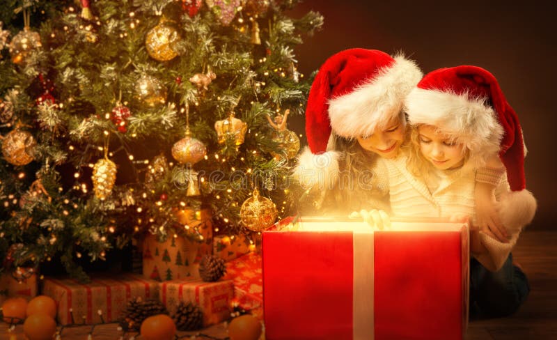 Christmas Children opening shining Red Present. Kids in Santa Hat looking at Light inside Gift Box next to decorated Xmas Tree