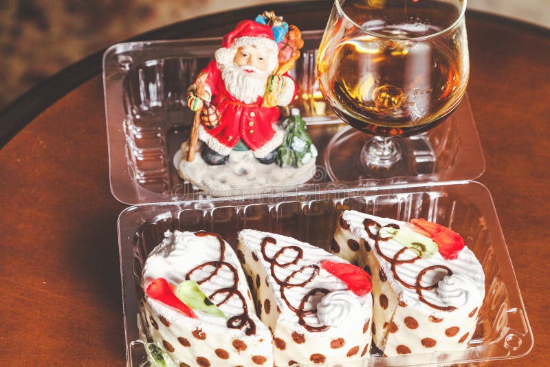 Christmas dessert set. Three mouth-watering cakes in the transparent box, brandy in elegant glass and the figure of Santa Claus in the background. Christmas dessert set. Three mouth-watering cakes in the transparent box, brandy in elegant glass and the figure of Santa Claus in the background