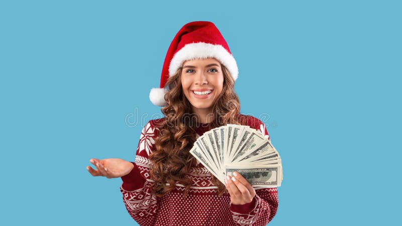 Christmas discounts concept. Happy millennial girl in Santa hat and sweater holding fan of money on blue background