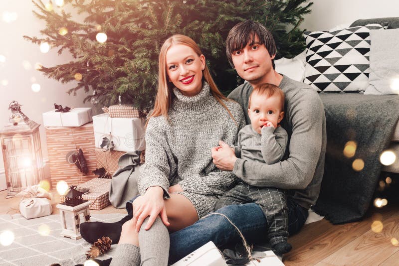 Christmas family with baby smiling near the Xmas tree. Living room decorated by Christmas tree and present gift boxes, the light royalty free stock photo Christmas family with baby smiling near the Xmas tree. Living room decorated by Christmas tree and present gift boxes, the light royalty free stock photo