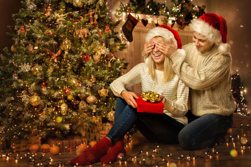 Christmas Family Couple with Gift. Man giving Present to surprised Woman next to Xmas tree. Closing eyes to Girlfriend in Room. Christmas Family Couple with Gift