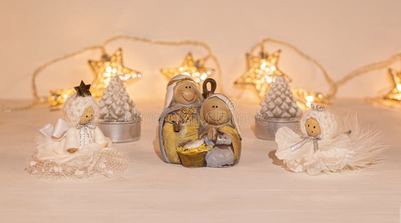Christmas festive scenery showing the Holy Family with 2 handcrafted angel ornaments in warm golden light; front view with blurred bokeh star lights background on white wooden table