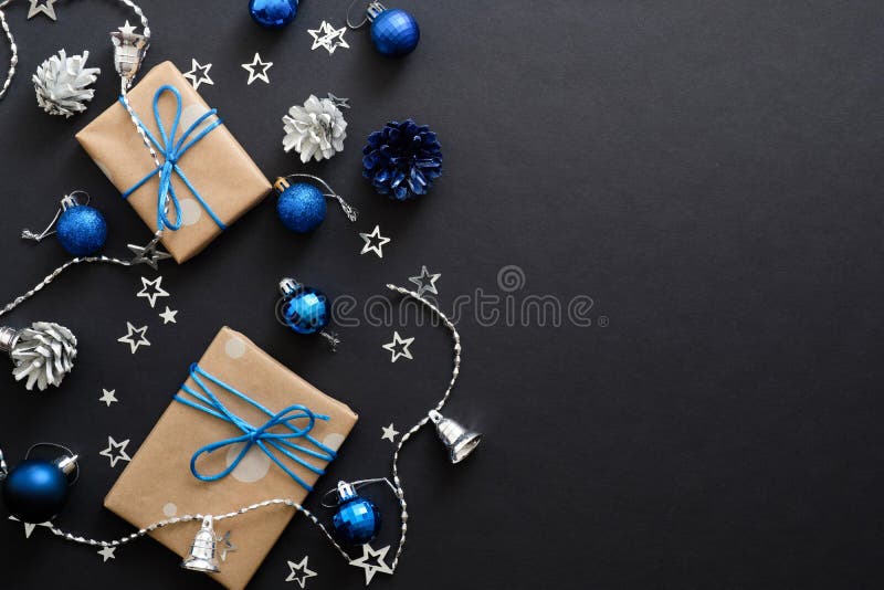 Christmas gifts with blue ribbon, blue balls, fir tree branches, cones, confetti on dark black background with copy space. Flat