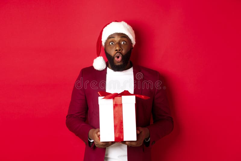 Christmas, New Year and shopping concept. Surprised african american man in santa hat saying wow, holding holiday gift