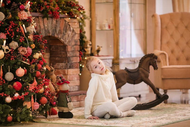 Christmas portrait of happy blonde child girl in white sweater siting on the floor near the Christmas tree and wooden toy horse. N stock photos Christmas portrait of happy blonde child girl in white sweater siting on the floor near the Christmas tree and wooden toy horse. N stock photos
