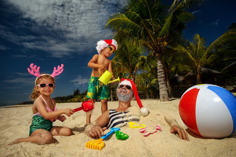 Christmas Santa Claus plays with kids. They are on tropical sandy beach with palm trees - Christmas travel discounts and travel